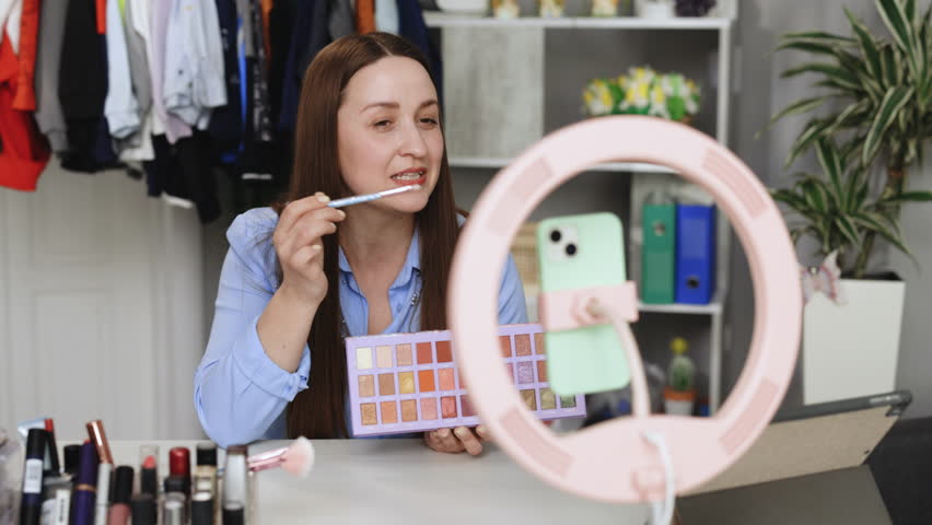 Young woman applying makeup on face with brush while recording vlog for social media via smartphone with tripod with LED ring lamp at table Royalty-Free Stock Footage #1104537789