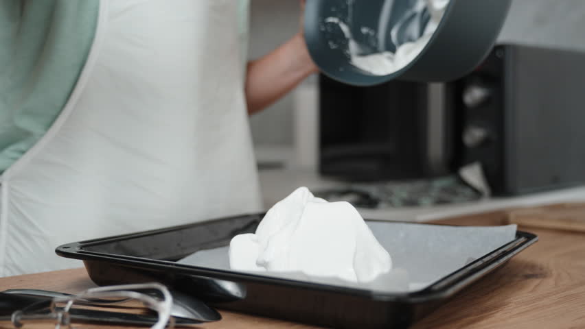 The pastry chef is piping meringue onto a baking sheet covered with parchment paper, in a close-up shot.
