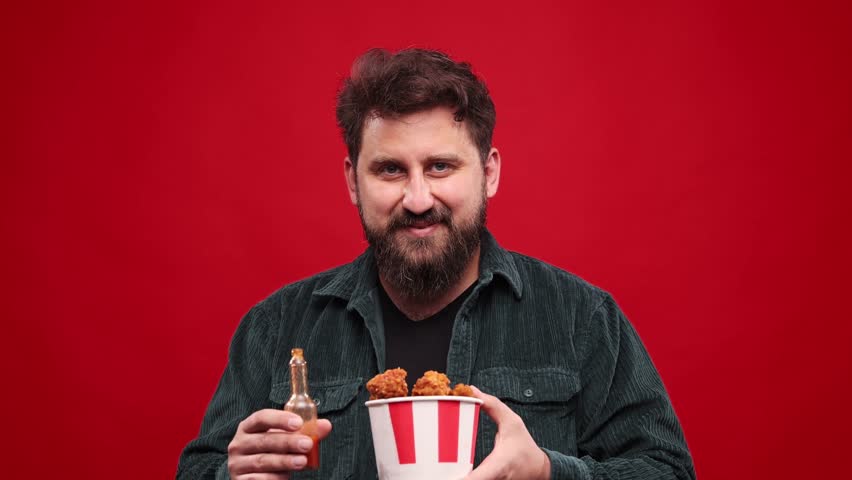 Funny crazy face. Bearded man eating fried chicken with hot chilli sauce against red studio background. Changing emotions, burning mouth. Spicy taste. Fast food lover. Concept of food, taste. Ad