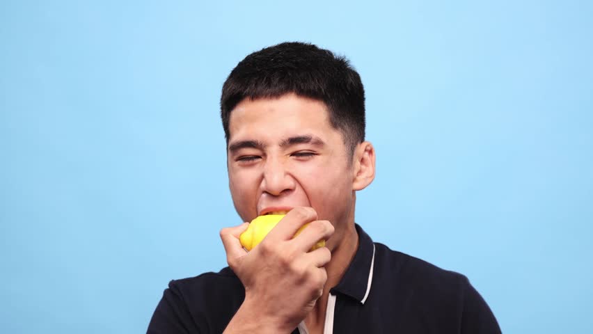 Young asian guy biting lemon against blue studio background. Handsome young man with grimacing face eating sour citrus. Concept of taste, human emotions, facial expression