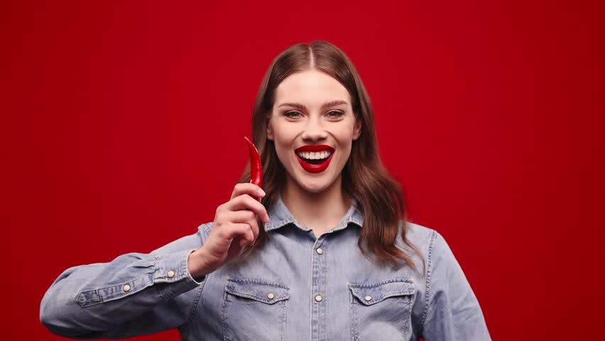 Changing emotions, Prety young girl with red lisptick eatingn red hot cilli pepers with smiling and grimacing face. Model agasint red background. Concept of food, taste, grocery, human emotions. Ad