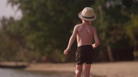 A video showing the back view of a little boy wearing a hat running in slow motion along the shore of a beach in Thailand during daytime