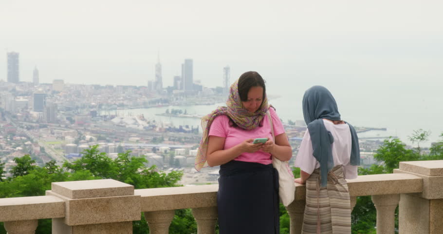 Mom daughter on terrace of Holy Trinity Church in headscarves in Georgia. Teen girl admiring amazing Batumi city and sea view from Sambeta Mountain. Woman browsing internet on smartphone. Tourism.