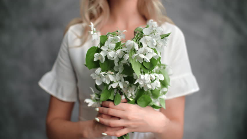Portrait unrecognizable blonde woman posing with gentle white flowers bouquet isolated on gray studio background closeup. Romantic young female holding spring blossom floral botanical natural bloom