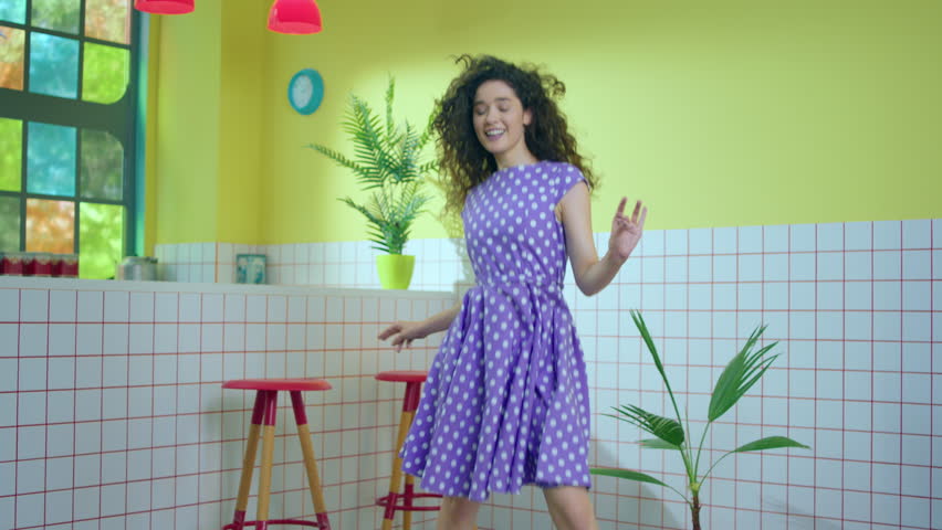 Amazing charismatic woman with a large smile moving very excited and dancing in front of the camera in the kitchen she wearing a beautiful pink dress