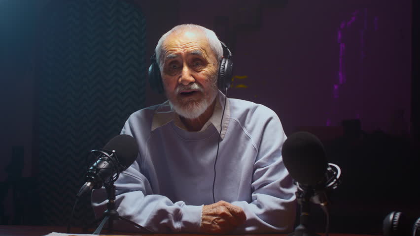 The old man creates a podcast in a professional studio. Senior male does online streaming interviews. Elderly man blogger speaking in microphone or make vlog. Recording audio content for social media