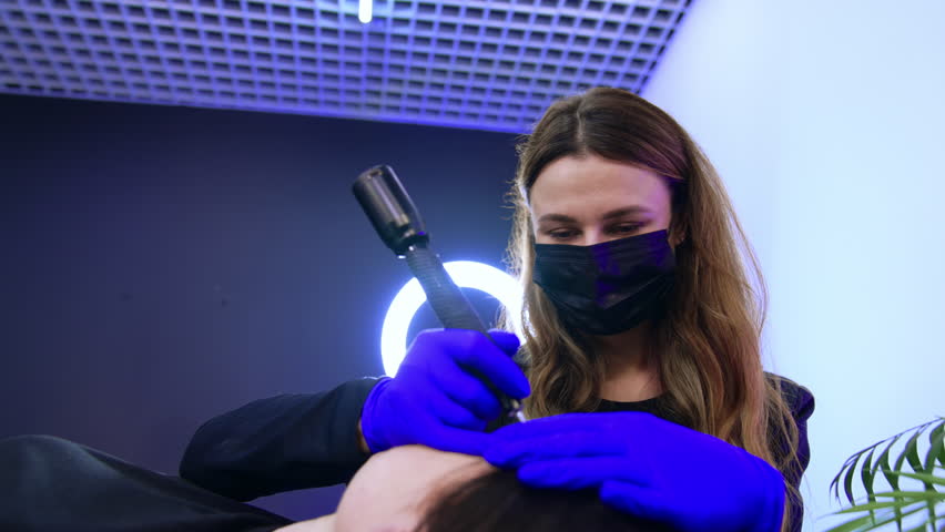 Long-haired beauty specialist in mask works in salon. Cosmetologist uses device to do the permanent eyebrow make-up. Low angle view. | Shutterstock HD Video #1104571149