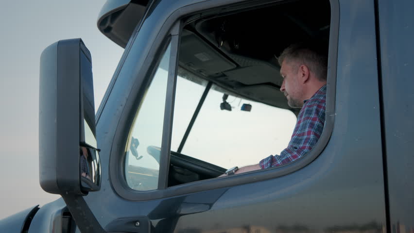 Professional Truck Driver sits in his truck and Behind Him Parked Long Haul Semi-Truck  Royalty-Free Stock Footage #1104572623