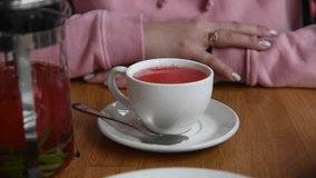 Close-up view of woman in pink sweatshirt stirring fruit tea with cowberry and rosemary in white cup using teaspoon in restaurant. Soft focus. Real time video. Healthy hot drinks theme.