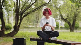 Young asian girl sitting on a bench outdoors in the city park, listening to music on headphones and smiling, typing message to friends on a smartphone. Concept of stylish youth who use gadgets - Powered by Shutterstock - Get 15% off with code: PIKWIZARD15