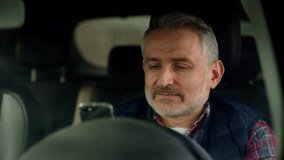 A man in a plaid shirt sits in a car and carefully looks at the smartphone screen. The driver of the car begins to rejoice after reading the good news on the smartphone.