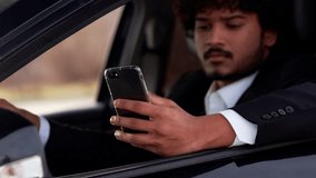 A gloomy young businessman sits in the car and taps on the smartphone screen. A man in a strict business suit drinks coffee while sitting in a car and looks at the smartphone screen.