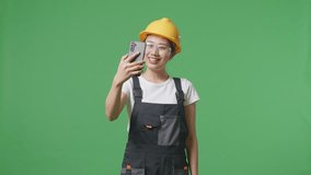 Asian Woman Worker Wearing Goggles And Safety Helmet Waving Hand Having A Video Call On Smartphone While Standing In The Green Screen Background Studio
