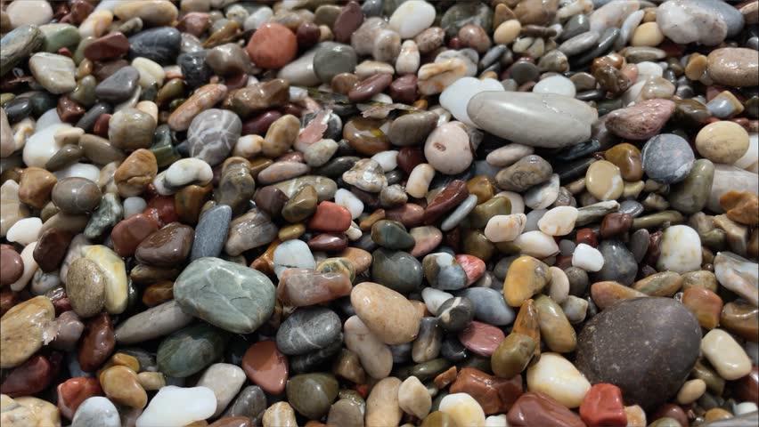 Closeup detail - small wet pebbles or sand on a beach, blurred afternoon sun lit calm sea in background, camera slides slowly to side. High quality 4k footage