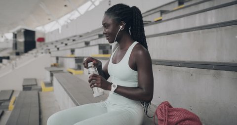 Black woman, music and drinking water in fitness arena stairs listening to workout playlist. Radio headphones, streaming audio and young African female person with podcast on stadium steps on break วิดีโอสต็อก