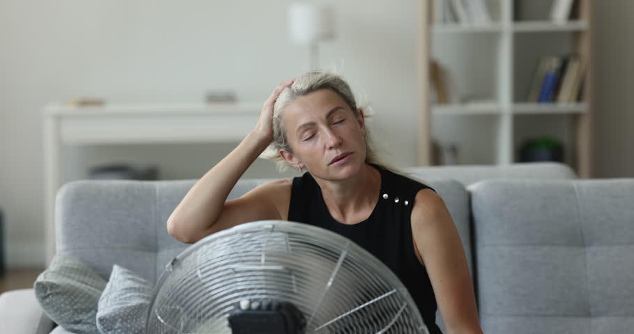 Exhausted mature homeowner woman getting cool, refresh at electric fan, suffering from overheating, hypoxia, sitting on sofa at fresh air blowing from propeller, feeling sick, tired, stressed Royalty-Free Stock Footage #1104601619