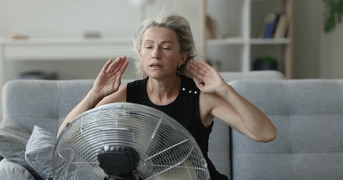 Exhausted mature homeowner woman getting cool, refresh at electric fan, suffering from overheating, hypoxia, sitting on sofa at fresh air blowing from propeller, feeling sick, tired, stressed Adlı Stok Video