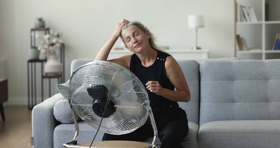 Positive peaceful mature senior woman relaxing at fresh cool air blowing from fan, sitting on home couch, enjoying freshness from blower, propeller, cooler, using domestic equipment for ventilation Royalty-Free Stock Footage #1104601651