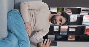 Vertical shoot. Portrait of bearded man sitting on sofa and holding digital gadget. Multicultural adult boy communicates with family via online call, gesturing and waving goodbye.