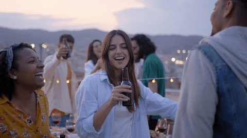 Three happy multiracial young people dancing at rooftop party with friends in background. Excited persons toasting with beer at barbecue in evening. Colleagues celebrating weekend break outdoors. Adlı Stok Video