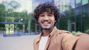 POV Close up Young cheerful smiling man talking on a video call using a smartphone while standing near an office building. Happy handsome male waving hand, speaks emotionally, laughs looking at camera
