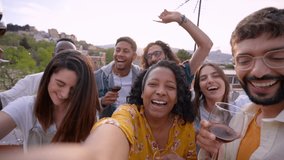 Point of view of cheerful young African American woman shooting video for blog about party with friends on rooftop. People looking at camera, dancing and holding wine glasses. Social networking.