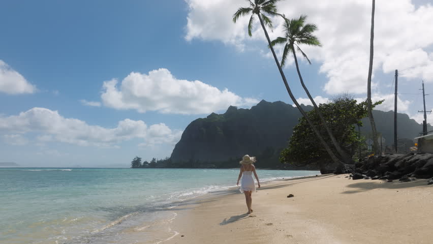 Slow motion Female tourist on summer vacation on paradise island of Oahu. Woman in white mini boho dress on green Hawaiian islands, USA 4K. Woman walking by tropical sandy beach leaving footprints Royalty-Free Stock Footage #1104613699