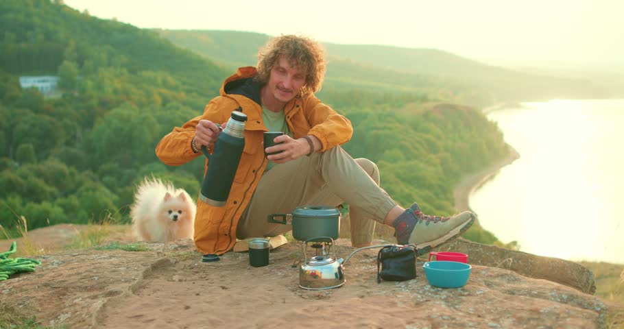 young man camper tourist pouring hot tea, going to drink coffee while sitting resting on the top peak of mountain, white Volpino Italiano Spitz pet walking behind owner, holiday adventure break