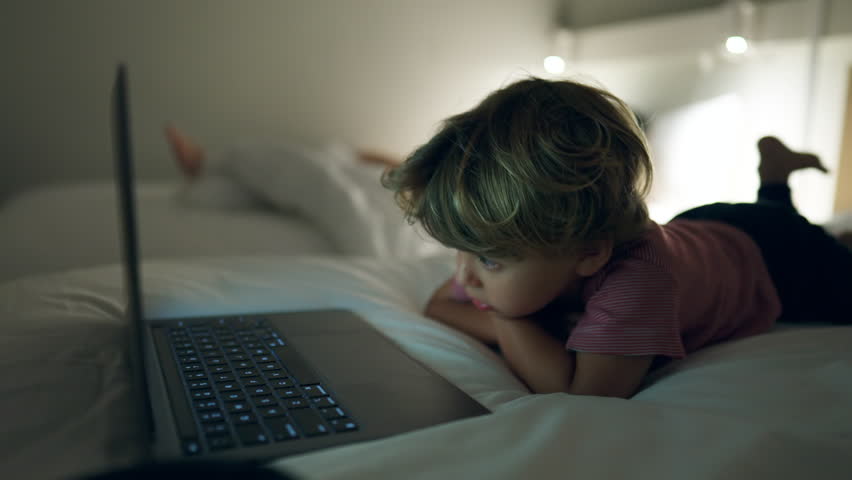 Small boy watching cartoons laying in bed at night. Child hypnotized by media online close up face in the evening