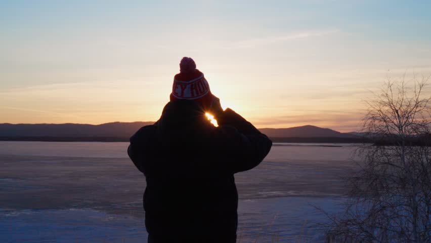 A young man stands on the shore of a frozen lake and looks through binoculars at the sunset. Travel and tourism.