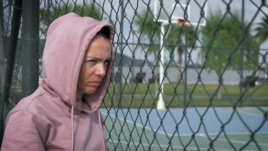 The female is in despair. A serious woman with a hood on her head stands sadly on the playground near the iron grid. The problem of assimilation of refugees. Royalty-Free Stock Footage #1104631611