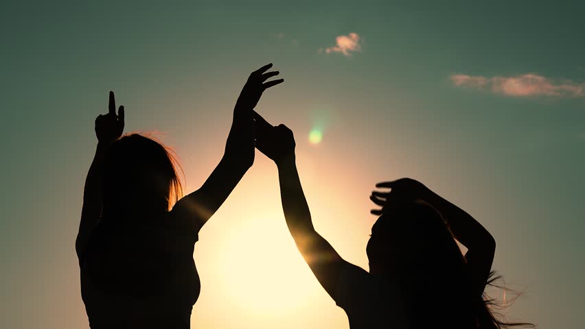 Friends have fun at beach party during sunset. Free girls dance merrily on summer evening, sun. Group of young people having fun dancing with raised hands to music. Celebrate summer day at beach party