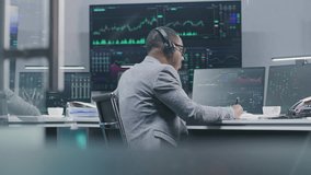 African American financial analyst monitors real-time stocks on computers, writes data with pen, works in bank office. Exchange market charts displayed on big digital screens. Investment or analytics. - Powered by Shutterstock - Get 15% off with code: PIKWIZARD15