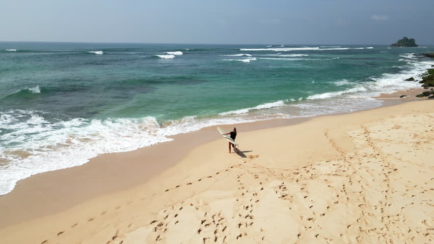 Surfer girl with surfboard walking on the beach towards ocean tide view from drone. Aerial shot of female surfer and shadow walking along the beach carrying longboard. Woman walking in tide.