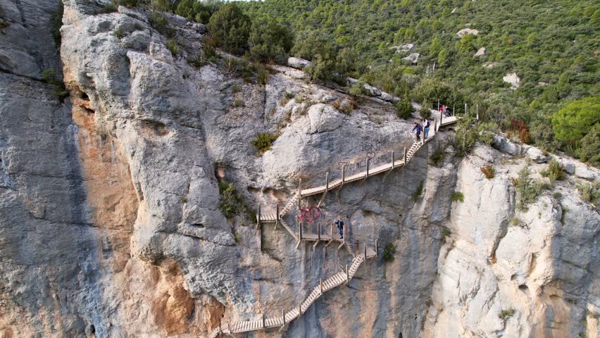 Panoramic beautiful vertiginous impressive aerial view of wooden staircase at rock cliff as part of hiking path in Congost de Montrebei gorge in Pyrenees, Spain. Group of people going up stairs. 4K.