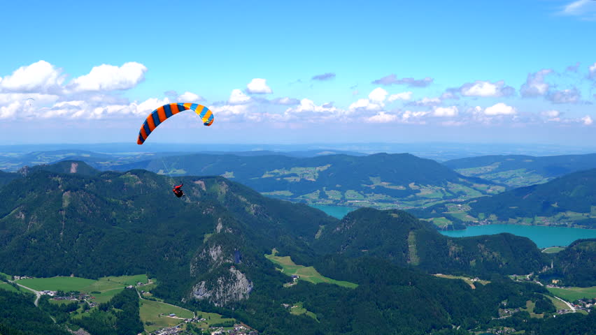 Paraglider flying over mountains and valleys on a summer, clear, day. Spectacular view of the Alps and Salzkammergut peak mountains and lake. Paragliding over a wild breathtaking nature with blue sky. Royalty-Free Stock Footage #1104640493