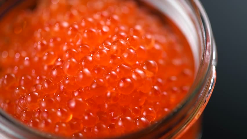 Red caviar in a glass jar, close up. Salmon caviar rotating over black background. Delicious gourmet food.  Delicatessen. Texture of caviar. Seafood, macro shot. 