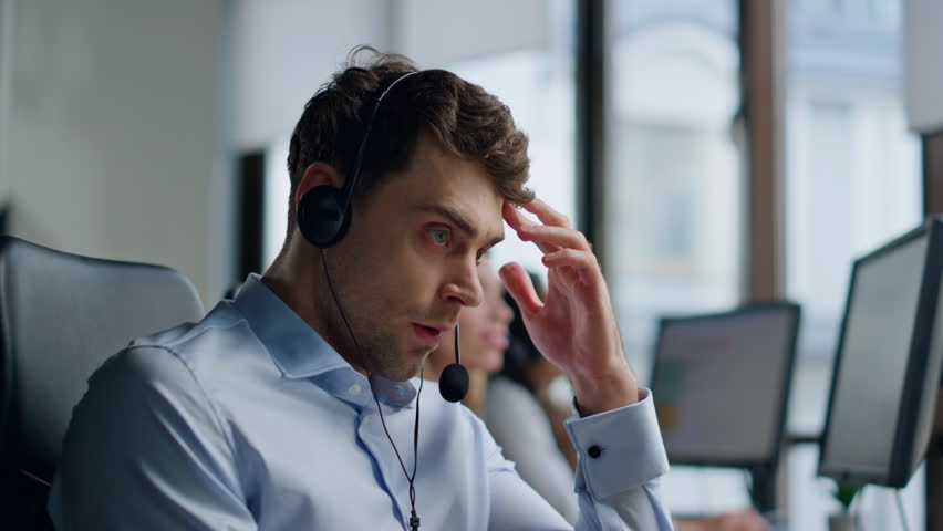Nervous man talking headset in call center. Stressed support agent work hotline. Annoyed focused specialist speaking customer explaining technical instructions. Thoughtful professional consult client Royalty-Free Stock Footage #1104643603