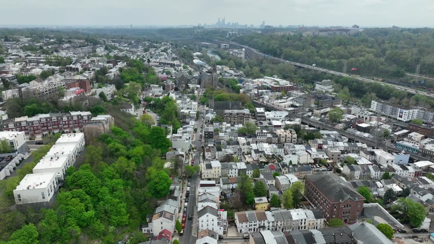 High aerial establishing shot of Manayunk, Pennsylvania. Philadelphia suburb with skyline in distance on cloudy day. Large town with many homes.