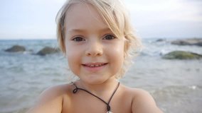 A cute little child video chats on smart phone on the beach on blue sea background. Using a portable handheld device on summer vacation. Talking on a mobile. Doing selfie. Wide angle view