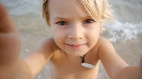 A cute little child video chats on smart phone on the beach on blue sea background. Using a portable handheld device on summer vacation. Talking on a mobile. Doing selfie. Wide angle view