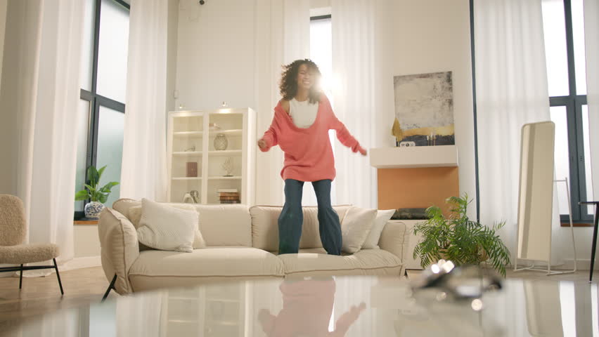 Afro girl homeowner having fun on move in day, celebrating mortgage approval, relocation to upgraded apartment. Bank mortgage concept. Excited happy young woman of color jumping happily on modern sofa Royalty-Free Stock Footage #1104654471