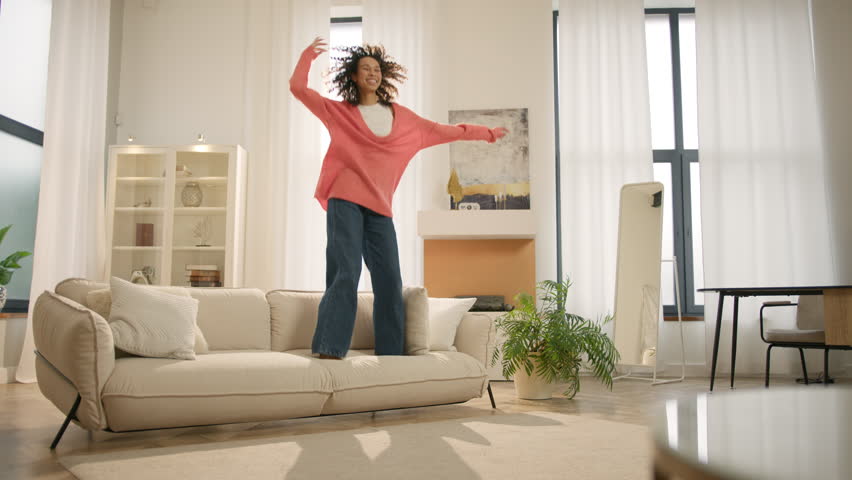 Afro girl homeowner having fun on move in day, celebrating mortgage approval, relocation to upgraded apartment. Bank mortgage concept. Excited happy young woman of color jumping happily on modern sofa