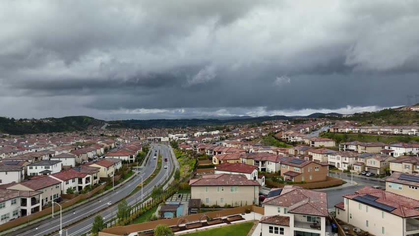 Aerial view of suburban tract homes in Santa Clarita, California on a rainy day with looming storm clouds overhead and wet roads. Many middle-income homes sit along the hillsides as traffic passes.