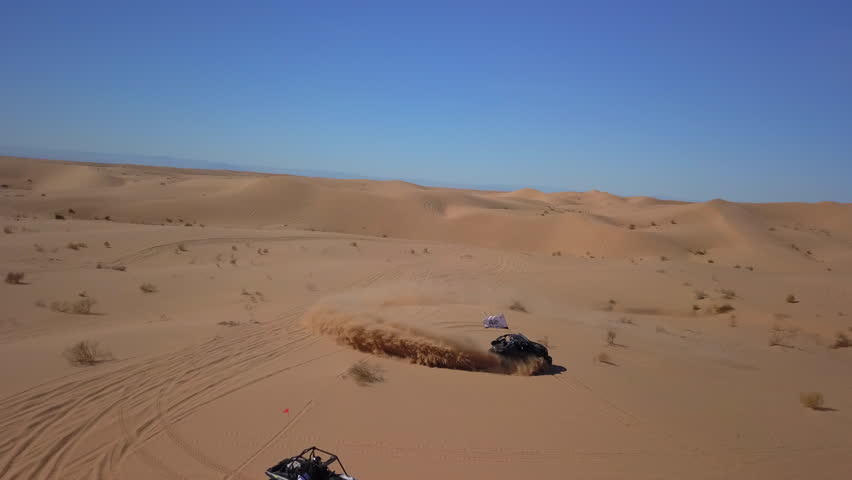 Aerial: Drone Flying Over Dune Buggy Doing Doughnut On Sand Amidst Bushes Against Blue Sky - Glamis, California Royalty-Free Stock Footage #1104664825