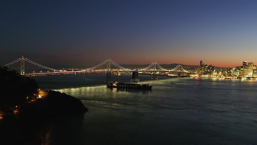 Aerial Shot Of Ship Moving Below Oakland Bay Bridge On Sea, Drone Descending Near Illuminated City Against Sky At Night - San Francisco, California