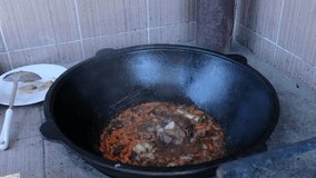 Woman's hand pours mixture of spices into boiling pilaf. Outdoor cooking in cauldron. Traditional lamb dish with fat tail fat. Mouth-watering video where traditional pilaf dish is cooked.