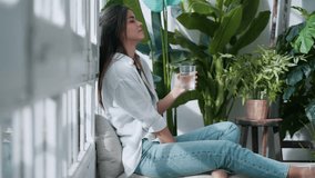 Video of young woman in lotus position drinking water while sitting on couch in the living room full of plants at home