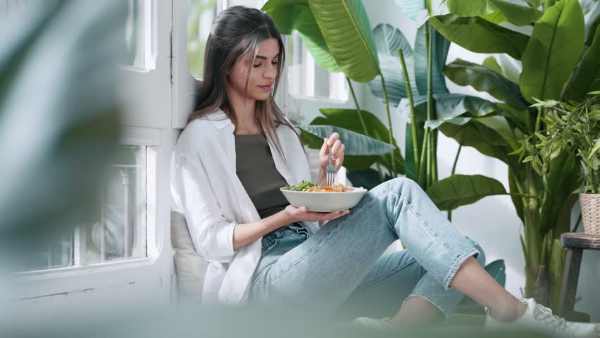 Video of young relaxing woman eating healthy salad while sitting on couch in the living room full of plants at home