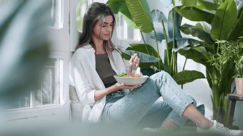 Video of young relaxing woman eating healthy salad while sitting on couch in the living room full of plants at home - Βίντεο στοκ